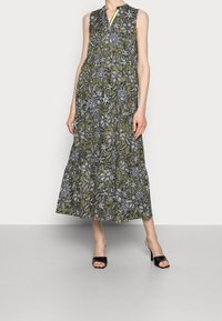 Vestido maxi sin mangas en verde oliva con patrones florales blancos. Presenta un cuello tipo camisero y un dobladillo en vuelos. Se combina con tacones negros de dedo abierto.