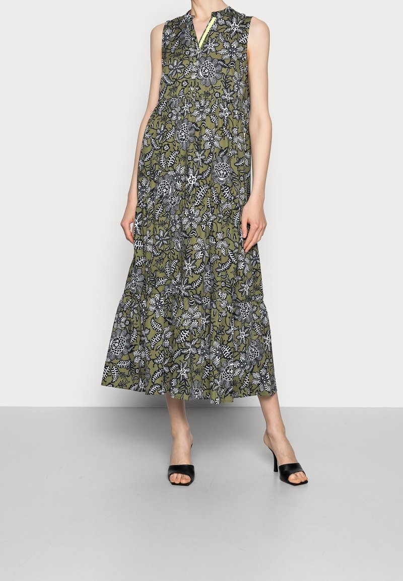 Vestido maxi sin mangas en verde oliva con patrones florales blancos. Presenta un cuello tipo camisero y un dobladillo en vuelos. Se combina con tacones negros de dedo abierto.