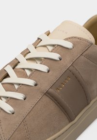 Κοντινό πλάνο μπεζ σουέντ αθλητικού παπουτσιού με λευκά κορδόνια, λείο δερμάτινο πάνελ και χρυσό λογότυπο "NUBIKK" στο πλάι.