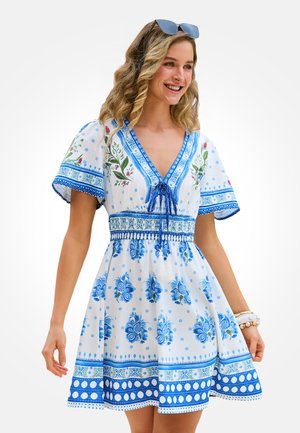 Uśmiechnięta kobieta w krótkiej sukience w biało-niebieskie kwiaty z bufkami na rękawach, dekoltem w serek i okularami przeciwsłonecznymi na głowie.