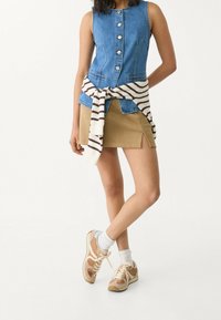 Denim ärmlös väst med knappstängning, i kombination med en beige minikjol med sidofickor. Vit och brun randig tröja knuten i midjan. Beiga sneakers.