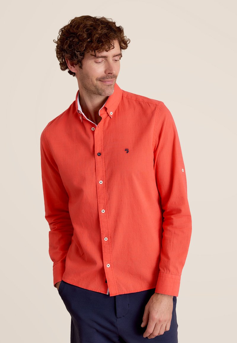 Chemise rouge à boutons avec une texture lisse, dotée d'un col blanc contrastant et d'un petit logo sur la poitrine, portée avec un pantalon bleu marine.