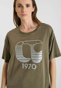 Olijfgroene crewneck t-shirt van zacht materiaal, met een grote zilveren grafische ontwerp met horizontale strepen en de tekst "1970".