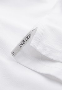 Gros plan d'un tissu blanc montrant une petite fente latérale avec une étiquette imprimée partiellement visible à l'intérieur de la couture.