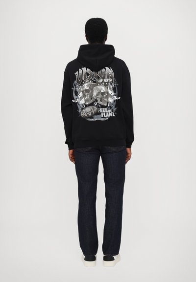 Haut noir à capuche avec un design graphique comportant deux crânes, du texte et des accents au dos. Associé à un jean vif foncé et des baskets blanches.