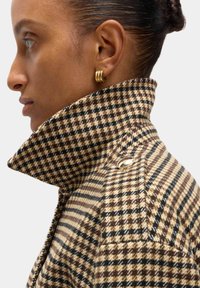 Bruin en zwart houndstooth patroon wollen jas met een hoge kraag, voorzien van een zilveren knoopaccent nabij de schouder.