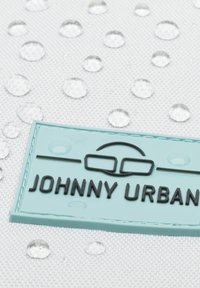 Étiquette en caoutchouc bleu clair avec le texte noir "JOHNNY URBAN" et un emblème de lunettes, sur un fond textile blanc avec des gouttes d'eau.