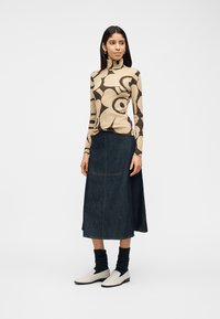 Γυναίκα που φοράει μπεζ πουλόβερ με λαιμόκοψη και μεγάλα σκούρα καφέ λουλουδένια σχέδια, μια σκούρα μπλε φούστα μήκους μεσαίου ύψους, μαύρες κάλτσες και λευκά μοκασίνια, στέκεται μπροστά σε λευκό φόντο.