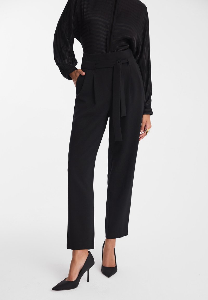 Czarne dopasowane spodnie wykonane z gładkiego materiału, z paskiem w talii, szerokimi nogawkami i bocznymi kieszeniami. Noszone z czarnymi szpilkami o spiczastym zakończeniu.
