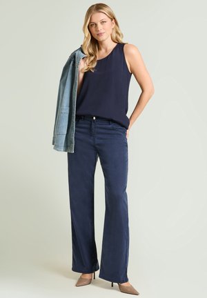 Vrouw met lang blond haar die een mouwloze marineblauwe top draagt, wijde marineblauwe broek, beige hakken, met een spijkerjasje over één schouder.