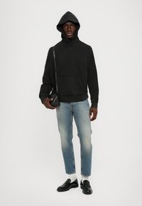 Schwarzer Hoodie mit einer Fronttasche, lockere Passform, kombiniert mit hellblauen Jeans, weißen Socken und schwarzen Loafers. Einfaches Design und lässiges Erscheinungsbild.