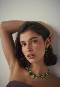 Grüne Edelsteinkette mit unterschiedlichen Formen, ergänzt durch bernsteinfarbene Akzente und eine goldene Kette; kombiniert mit passendem Ohrschmuck. Der Hintergrund ist neutral.