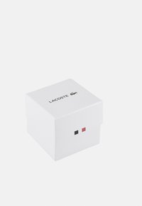 Cutie pătrată albă cu capac, având accente negre și roșii și logo-ul Lacoste imprimat pe partea superioară într-un font simplu.