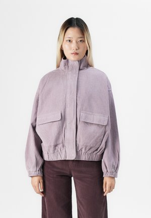 Young woman with straight blonde hair wears a lilac corduroy jacket with high collar and front pockets, paired with dark purple corduroy pants.