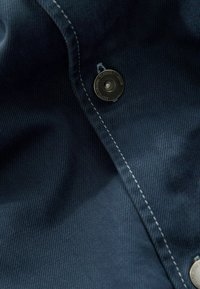 Tessuto blu navy con una trama testurizzata, dotato di un pulsante a scatto in metallo argentato e cuciture bianche a contrasto lungo il bordo.