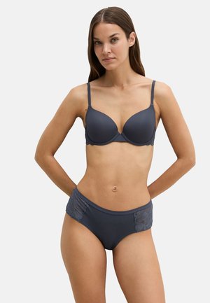 Vrouw met lang bruin haar die een bijpassende donkergrijze beha- en slipset met kanten details draagt, staat met handen achter haar rug.