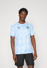 Šviesiai mėlynas futbolo marškin�ėliai su abstrakčiu raštu, trumpomis rankovėmis, apvalia iškirpte ir juodu Puma logotipu. Dėvimi su juodomis šortais.