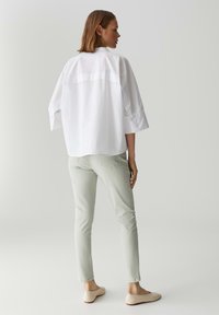 Camicia bianca oversize con maniche ampie, abbinata a pantaloni aderenti verde chiaro e ballerine beige. Tessuto testurizzato e dettagli di design sottili visibili.