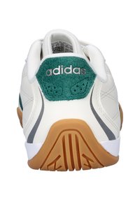 Zapatilla deportiva de cuero blanco con detalle de talón en ante verde, rayas grises y suela de goma color caramelo. Lleva la marca Adidas en la parte trasera.