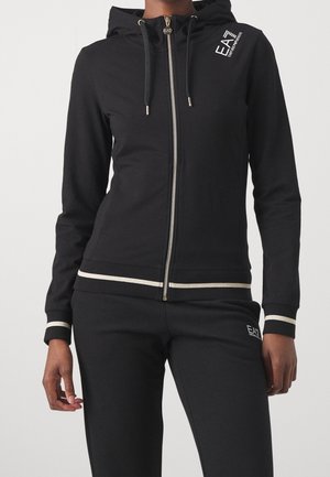 Personne portant un sweat à capuche noir zippé et un pantalon assorti avec des finitions blanches et des logos EA7 Emporio Armani sur la poitrine et la cuisse.