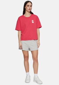 Rød bomulds t-shirt med et logo på brystet, kombineret med grå shorts. Outfittet har en afslappet pasform og er suppleret med hvide sneakers.