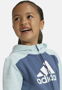 Sudadera con cremallera en azul y azul claro con un logo de Adidas en blanco. Presenta un diseño de color dividido, una textura suave y una capucha.