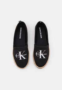 Des chaussures slip-on noires en toile avec une semelle en jute. Présentent un logo "CK" blanc bien visible et la mention "CALVIN KLEIN JEANS" imprimée à l'intérieur.