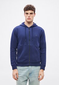 Tengerészkék cipzáras kapucnis pulóver puha anyagból, kapucnival, elülső zsebekkel és egy kis logófolttal a mellkason. Világoskék farmernadrággal párosítva.