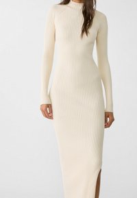Elfenbeinfarbiges, geripptes Strickkleid mit langen Ärmeln, hohem Ausschnitt und seitlichem Schlitz, das eine figurbetonte Silhouette und strukturierte Stoffe aufweist.