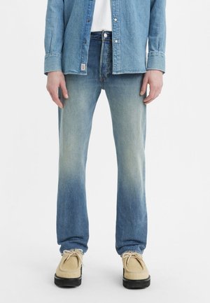 Jean en denim bleu clair avec un dégradé délavé, associée à une chemise en chambray bleu clair et des chaussures beiges, affichant un look décontracté.
