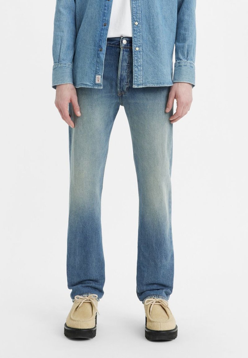 Světle modré džíny z denimu s vybledlým přechodem, spárované se světle modrou košilí z chambray a béžovými botami, které vykreslují ležérní vzhled.