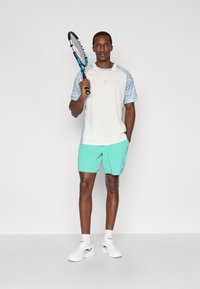 Τένις ντύσιμο με ανοιχτό γκρι πουκάμισο με μπλε μανίκια με σχέδια, σορτς μέντας και λευκά παπούτσια τένις, κρατώντας ρακέτα.