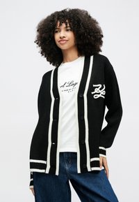 Kobieta o kręconych włosach, ubrana w czarno-biały kardigan na białej koszulce z grafiką i niebieskich dżinsach, stoi na jednolitym tle.