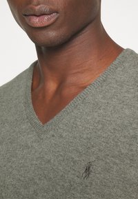 Grå stickad v-ringad tröja med en texturerad yta och en liten svart broderad logotyp på vänstra bröstet.