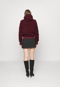 Burgundská fleece mikina s vysokým límcem a strukturovanou tkaninou, spárovaná s černou mini sukni a černými vysokými botami.