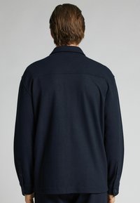 Chaqueta textura en azul marino, con cuello tipo solapa, puños abotonados y un corte relajado. Presenta costuras simples y un tejido ligero.