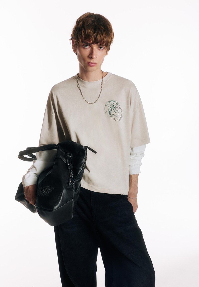 Jeune personne aux cheveux courts et bruns portant des chemises superposées beige et blanche, un pantalon noir, un collier chaîne, tenant un grand sac en cuir noir.