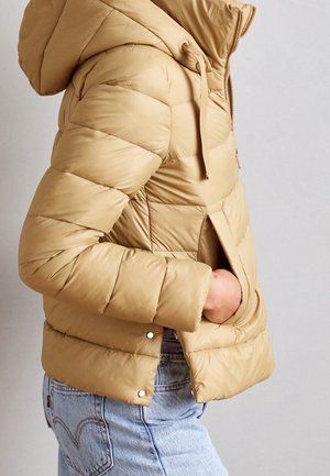 Osoba ubrana w beżową kurtkę pikowaną z kapturem, z ręką w bocznej kieszeni, w połączeniu z jasnoniebieskimi dżinsami na jednolitym tle.