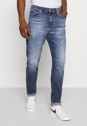 Mand iført falmede blå jeans med opsmøgede bukseben og hvide sneakers med burgundy Nike swoosh, hænderne i lommerne, stående mod en hvid baggrund.