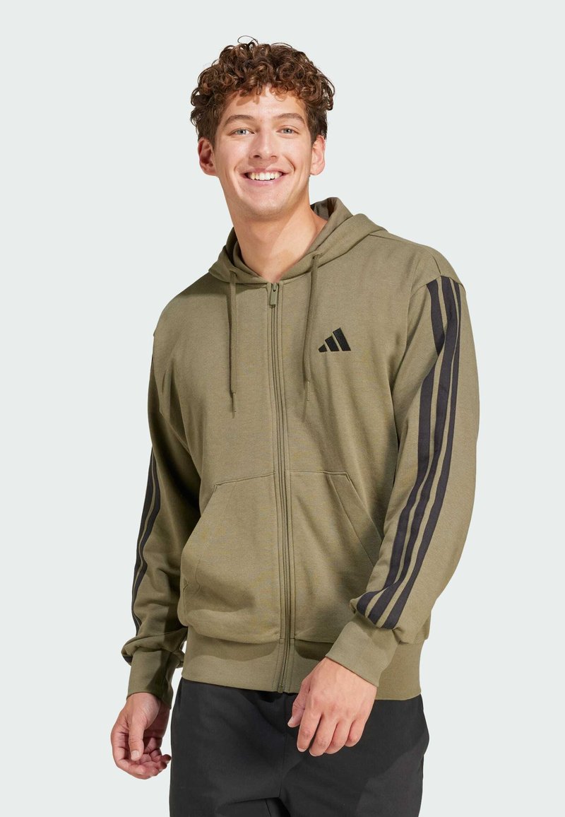 Veste à zip verte avec des rayures noires sur les manches et un petit logo noir sur la poitrine, fabriquée en tissu doux et texturé.