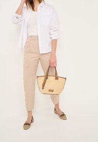 Sac à main beige avec des accents marron, texture tissée et une forme structurée. Le modèle porte une chemise blanche et un pantalon beige avec des mocassins à motifs.