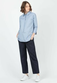 Camisa de manga larga azul claro con cuello abotonado, dobladillo redondeado y pliegue delantero. Combinada con pantalones oscuros de pierna recta de mezclilla y zapatillas blancas.
