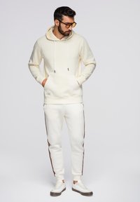 Crème hoodie met trekkoorden, voorzak; bijpassende crème joggingbroek met bruine zijkanten, gecombineerd met twee kleuren sneakers.
