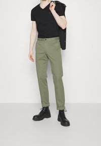 T-shirt nera a scollo a V, pantaloni verde oliva con passanti per cintura e tasche frontali, e stivali neri stringati, modello che tiene una giacca sulla spalla.