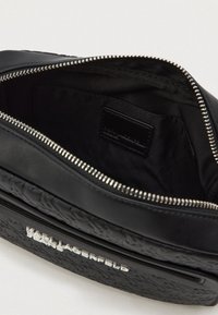 Bolsa em couro sintético preto com uma superfície texturizada, fecho prata e um interior elegante. Apresenta um rótulo interno e um logótipo da marca em relevo.