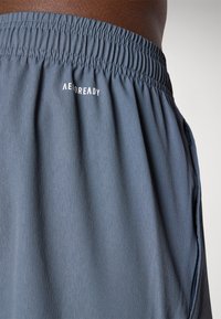 Et par blå træningsshorts med en elastisk talje og en sidelomme med lynlås. Stoffet har en subtil tekstur. Logoet "AEROREADY" er synligt.