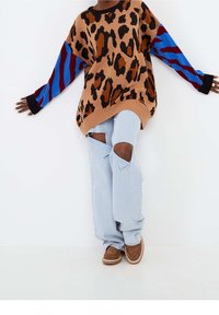 Personne portant un pull oversize à imprimé léopard avec des manches rayées bleues et bordeaux, un jean clair déchiré, et des chaussures marron.