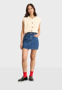 Top senza maniche a coste beige con bottoni; gonna mini in denim blu con tasche; scarpe nere; calze rosse. Modello in piedi davanti a uno sfondo grigio.