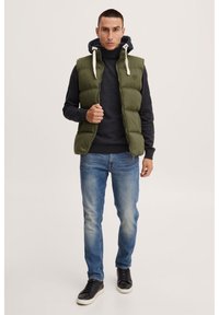 Grønn puffervest med høy krage, beige snor og emblem. Brukt over mørk genser og blå jeans, sammen med svarte sneakers.