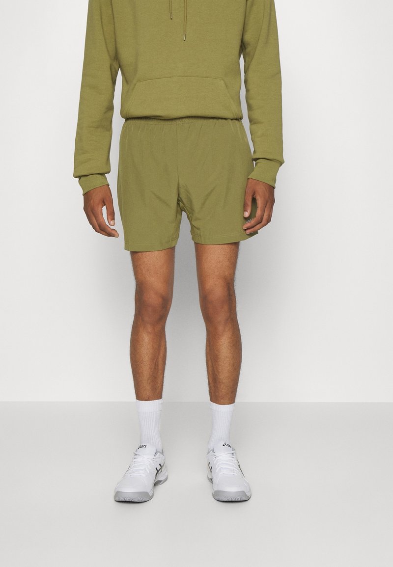 Sweat à capuche vert olive avec une poche avant et un cordon de serrage, associé à un short assorti. Des chaussettes blanches jusqu'aux chevilles et des chaussures de sport complètent la tenue.